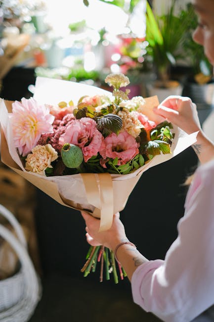 A woman in a red dress smiling as she receives a bouquet of white and green flowers from a delivery person wearing a high-visibility vest and red cap, standing outdoors near a brick wall and leafy gre