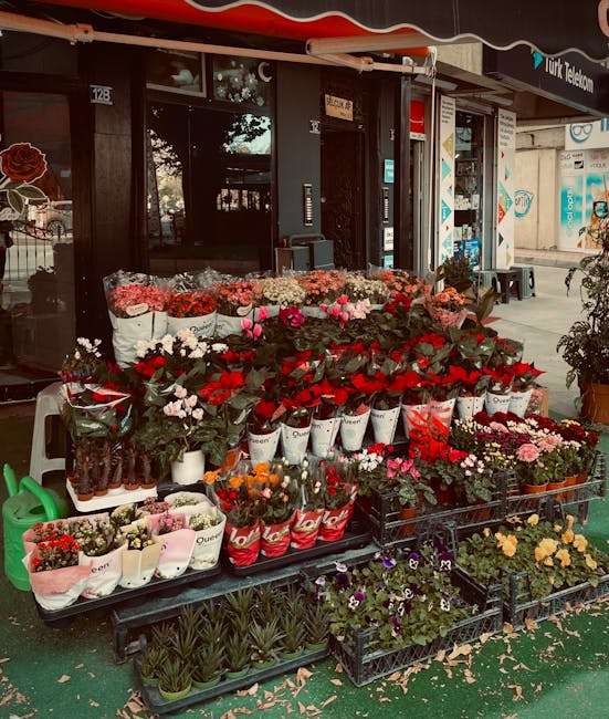 A vibrant display of fresh red and white flowers, including roses, carnations, and roses in heart-shaped balloons, arranged in baskets and wrapped in pink, red, and white paper. The bouquet includes g